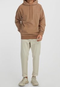Uomo che indossa un hoodie marrone, pantaloni beige con risvolto e sneakers beige, in piedi davanti a uno sfondo bianco semplice.