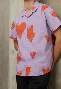 Košile s krátkým rukávem světle fialové barvy s červenými srdíčkovými vzory, s kapsou a zapínáním na knoflíky. Vyrobena z měkké látky.