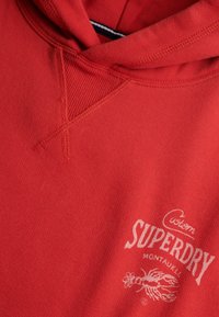 Roter Hoodie mit geripptem, V-förmigem Steppdetail und weißem "Custom Superdry Montauk L.I." Text über einer Hummergrafik auf der Brust.