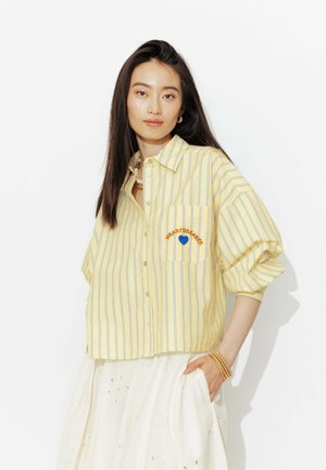 Femme aux longs cheveux foncés portant une chemise rayée jaune avec le texte « HEARTBREAKER » et un cœur bleu sur la poche, associée à une jupe blanche.