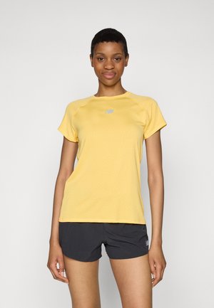 Ung kvinde iført en gul kortærmet sports skjorte og sorte shorts, stående og vendt fremad mod en ensfarvet baggrund.