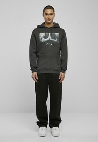 Dark grey hoodie with a front graphic of clasped hands, featuring white text below. Worn with loose black pants and white sneakers.
