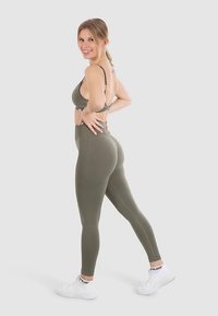 Olijfgroene sportbeha en leggings met hoge taille, beide met een gladde textuur. Witte sportschoenen met logo-accent, model poseert met handen in de zij.