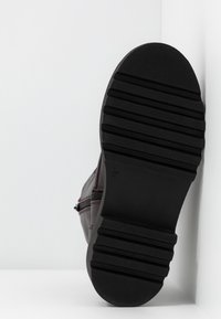 Semelle d'une botte brun foncé avec une épaisse semelle en caoutchouc noire présentant des rainures horizontales, inclinée contre un mur blanc d'angle et un sol.