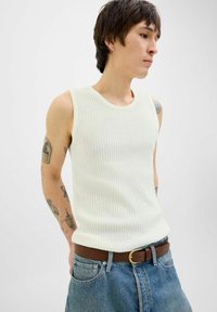 Geribbelde, mouwloze tanktop in lichtcrème kleur, gecombineerd met hooglopende blauwe jeans en een bruine riem. Zichtbare tatoeages op de arm.