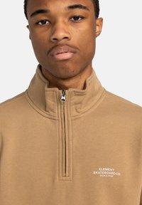 Pullover beige con design a zip a un quarto, texture morbida e logo bianco "Element Skateboard Co." sul petto.