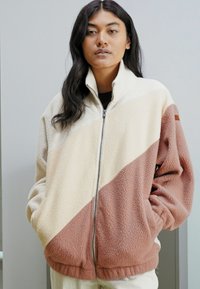Chaqueta de forro polar con cremallera frontal; presenta bloques de color crema, beige y óxido, cuello alto y bolsillos laterales. Textura suave, ajuste relajado.