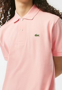 Polo en coton rose doux avec un col classique, des manches courtes et un logo crocodile brodé vert sur la poitrine gauche.