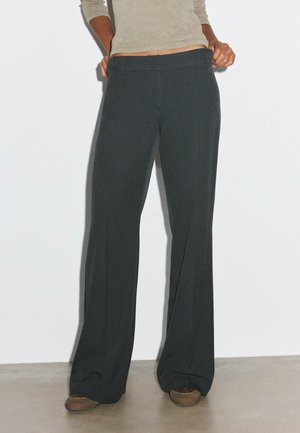 Personne portant un pantalon ample gris foncé et des chaussures marron, debout contre un mur blanc uni.