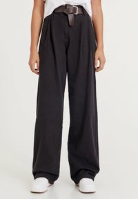 Czarne spodnie z szerokimi nogawkami i plisami, z brązowym skórzanym paskiem i białymi sneakersami. Wykonane z miękkiego, lekkiego materiału.