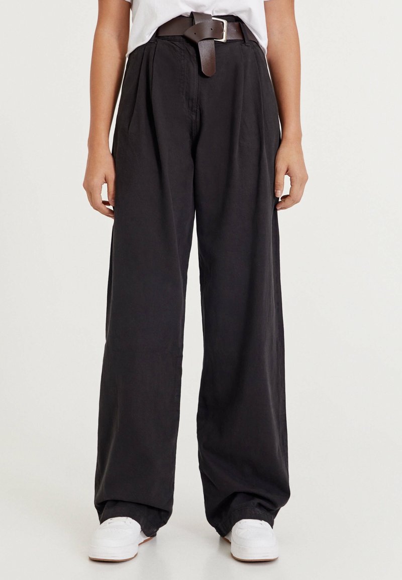 Czarne spodnie z szerokimi nogawkami i plisami, z brązowym skórzanym paskiem i białymi sneakersami. Wykonane z miękkiego, lekkiego materiału.