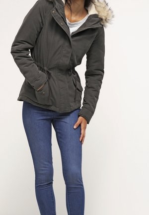 Vrouw in een donkergroene parka met capuchon en bontkraag, witte blouse en blauwe skinny jeans, staand tegen een effen achtergrond.