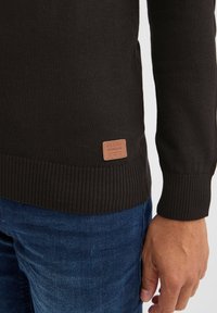 Zwarte gebreide trui met geribbelde manchetten en onderrand. Voorzien van een tan leren label met reliëftekst linksonder. Gecombineerd met blauwe jeans.