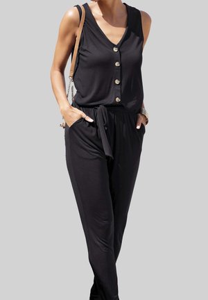 Kvinde iført sort ærmeløs buksedragt med knapper og sidelommer, bærende en lysebrun skuldertaske og iført armbånd.