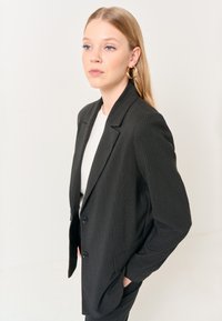 Blazer nero a righe con due bottoni, revers a punta, tasche laterali e un tessuto texturizzato. Indossato sopra una maglietta di colore chiaro.