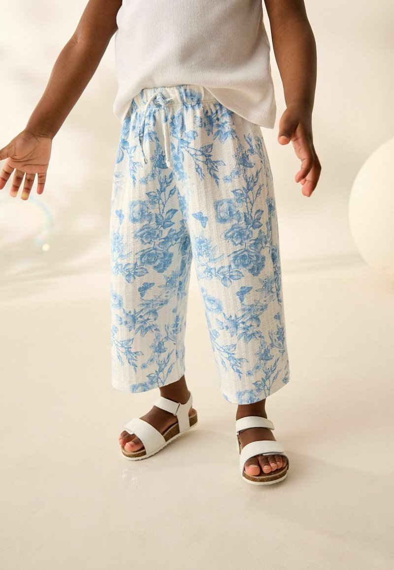 Kind trägt weiße Sandalen und lockere weiße Hose mit blauem Blumendruck, steht auf einer glatten hellen Oberfläche.