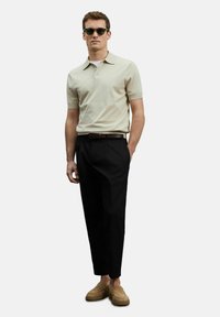 Beige polo shirt met korte mouwen, zwarte broek en bruin suède loafers. Model draagt zwarte zonnebril en presenteert een gepolijste look.