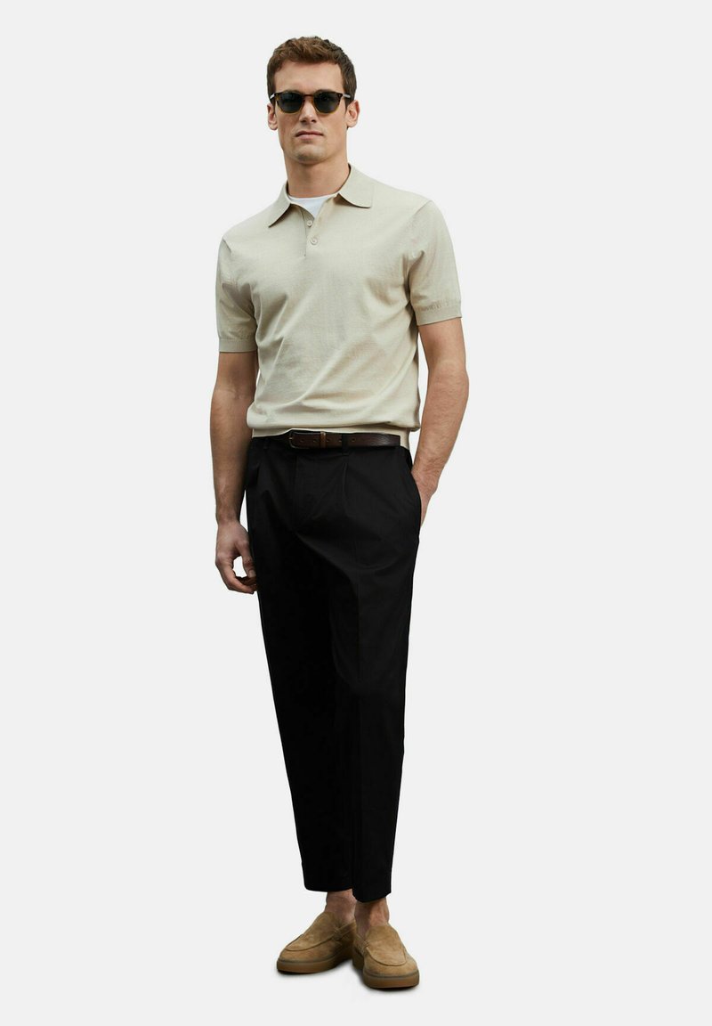 Beige polo shirt met korte mouwen, zwarte broek en bruin suède loafers. Model draagt zwarte zonnebril en presenteert een gepolijste look.