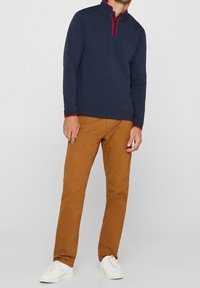 Marineblauer Pullover mit langen Ärmeln, roten Akzenten und Knopfleiste, kombiniert mit hellbraunen Slim-Fit-Hosen und weißen Sneakers. Glatte Baumwolltextur.