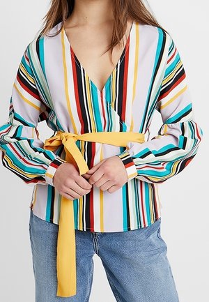 Persona con camiseta de manga larga y escote en V con rayas verticales de colores y un cinturón amarillo anudado en la cintura, combinado con vaqueros azul claro.