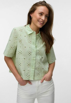 Femme souriante aux longs cheveux bruns porte un chemisier vert clair à manches courtes avec des motifs floraux en découpe et un pantalon blanc, les mains dans les poches.