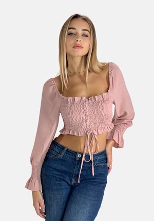 Haut court rose, avec des détails smockés, un décolleté carré et de longues manches bouffantes. Associé à un jean taille haute bleu.