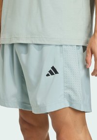 Pantalones cortos deportivos azul claro con logo negro de Adidas, con detalles perforados y textura suave, combinados con una camiseta gris.