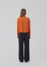 Chemise orange raccourcie à manches longues avec poignets boutonnés, associée à un pantalon noir à jambes larges. Texture lisse, design simple, sans motifs.