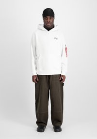 Un sweat à capuche blanc avec poche, logo étoile brodé, associé à un pantalon cargo olive et des chaussures noires à enfiler. Un couvre-chef noir complète la tenue.