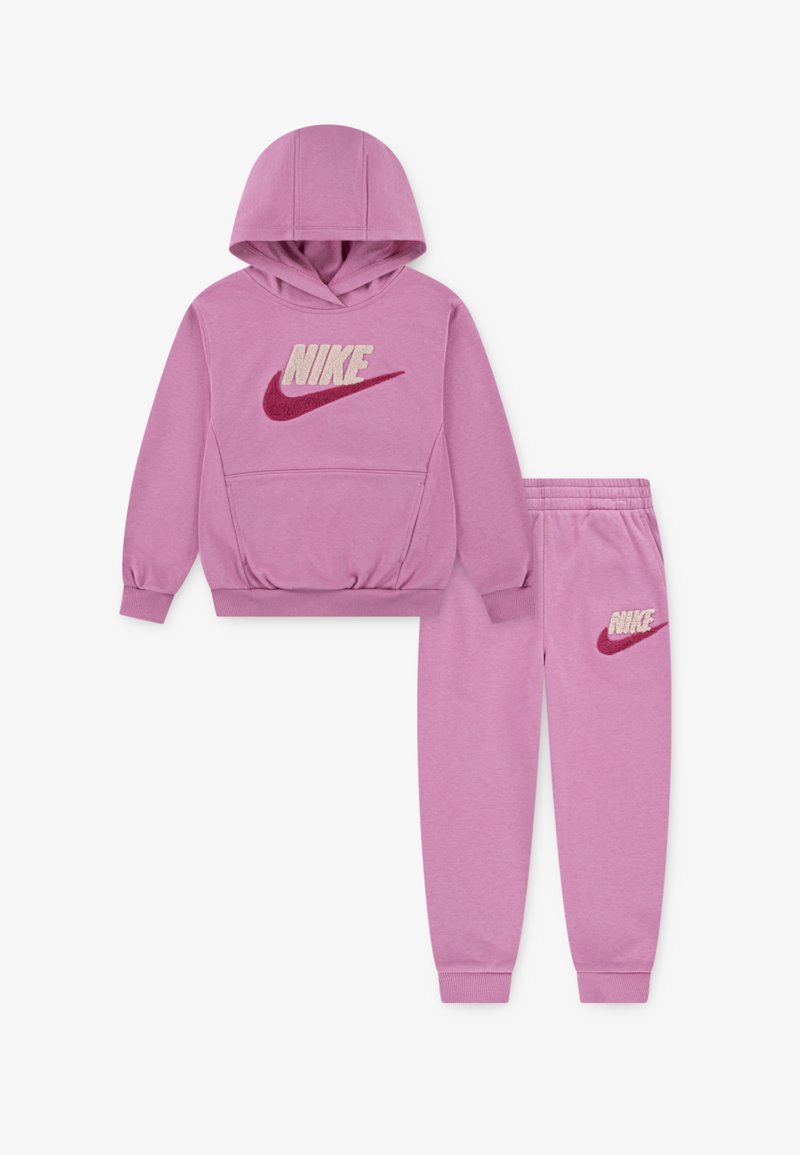 Różowy zestaw bluzy i spodni dresowych wykonany z miękkiego materiału. Bluza ma duże, puszyste logo Nike; spodnie mają mniejsze logo na udzie.
