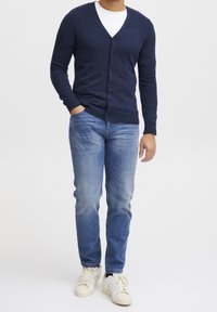 Navy v-neck kardigan dengan detail bahu bertekstur ribbed, depan berbutton, dipasangkan dengan celana jeans biru muda dan sepatu sneakers putih. Tekstur kain yang halus.