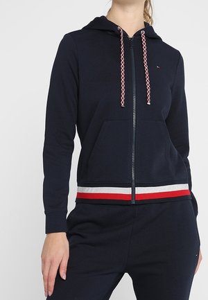 Woman wearing a navy zip-up hoodie with red, white, and gray striped trim and matching navy pants.