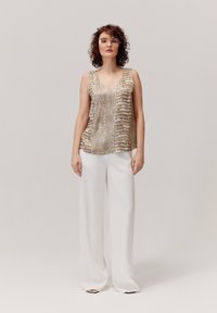 Metaliczna złota bluzka bez rękawów z dekoltem w serek i wzorem krokodyla, zestawiona z białymi spodniami typu wide-leg. Gładka tekstura, minimalistyczny design.