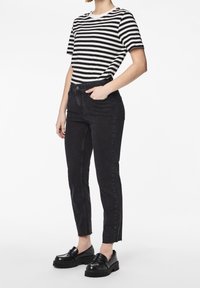 Persona con jeans negros de tiro alto, mocasines negros gruesos y camiseta de mangas cortas a rayas negras y blancas, con la mano en el bolsillo, de pie.