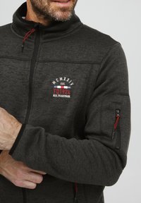 Mørkegrå fleecejakke med glidelås, høy krage og rød snor. Har brodert logo og sidelomme med glidelås.