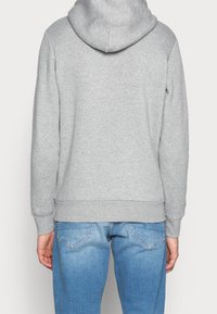 Persona che indossa una felpa con cappuccio grigia chiara e jeans blu, vista di spalle su uno sfondo semplice.