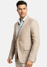 Blazer de lino beige con dos botones, bolsillos laterales y un acabado texturizado. Lleva puesto sobre una camisa azul clara con un patrón floral.