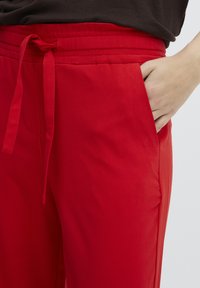 Red trousers made of lightweight fabric, featuring a drawstring waist and side pocket. Smooth texture with a loose fit.