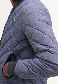 Prošívaná modrá bomber bunda, navržená s texturovanou tkaninou, žebrovanými manžetami a malým logem na hrudi. Obsahuje boční kapsy.