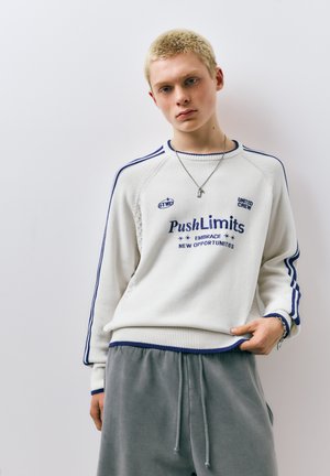 Ung person med kort blondt hår iført en hvid sweater med blå striber og tekst, grå bukser og en halskæde med nøgler vedhæng, stående op ad en hvid væg.