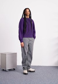 Purple sweatshirt with a crew neckline, paired with light gray pants. Model wears white shoes with dark soles. Background is plain white.