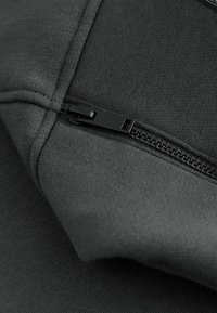 Giacchetto nero con zip, caratterizzato da un tessuto testurizzato e da un pratico tirante della zip, che evidenzia le cuciture e i denti in metallo nero della zip.