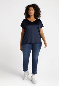Tricou V din material neted de culoare bleumarin, asortat cu blugi desenați în nuanțe de albastru, cu tivuri destrămate și detalii cu nasturi. Tenisi albi completează aspectul.