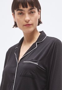 Mustad pidžaamaülemine valgete äärtega, ees nööpidega ja rinnataskuga. Pehme kangas, sileda tekstuuriga.