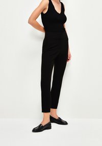 Zwarte getailleerde broek met een hoge taille, een gladde textuur en zijzakken. Draagt een zwart mouwloos topje en zwarte instappers.