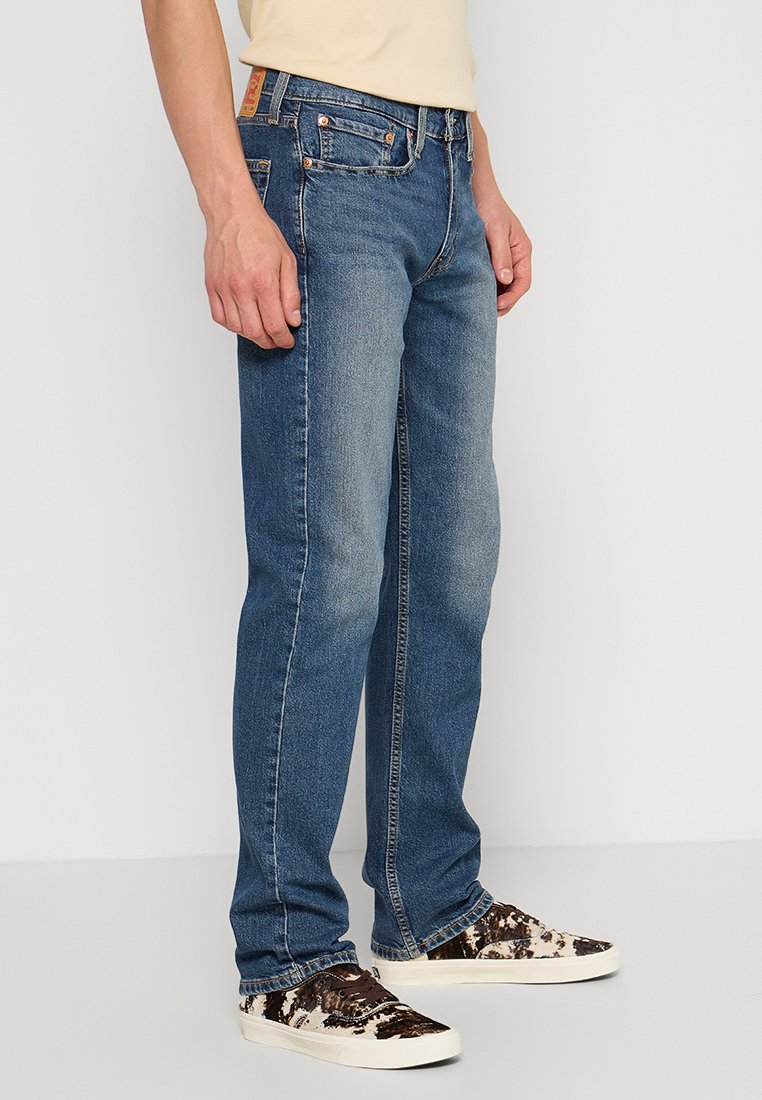 Man med raka blå jeans och bruna och vita mönstrade sneakers som står mot en vit vägg.