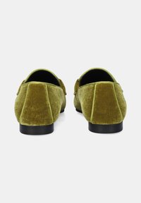 Un par de mocasines de terciopelo verde oliva con suelas negras vistos desde atrás sobre un fondo blanco.