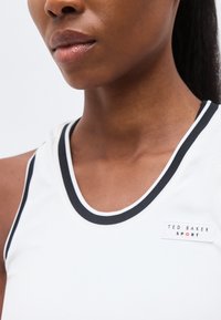 Κοντινό πλάνο ενός ατόμου που φοράει λευκή αμάνικη μπλούζα με μαύρα περιθώρια και ετικέτα "Ted Baker Sport" στο στήθος.