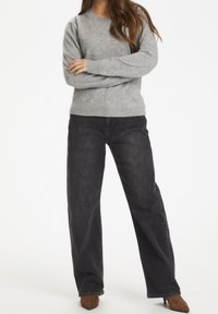 Femme debout, bras croisés, portant un pull gris, un jean noir à jambes larges et des bottines à talons pointues marron sur un fond blanc.