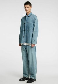 Lichtblauw denimjack met grote borstzak, knoopsluiting en losse pasvorm, gecombineerd met bijpassende losse denimbroek en zwarte schoenen.